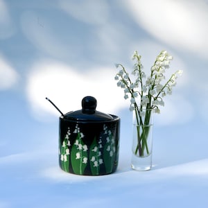 May include: A black ceramic sugar bowl with a lid and a small spoon, decorated with white lily of the valley flowers and green leaves. A small glass vase holds a bouquet of lily of the valley flowers.