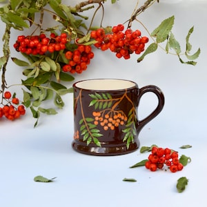 May include: A dark brown ceramic mug with a cream interior, decorated with a hand-painted design of orange berries and green leaves. The mug is set against a backdrop of similar berries and foliage.