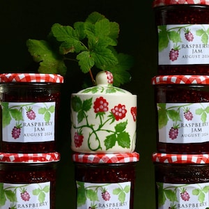 May include: Six jars of raspberry jam with red and white gingham lids. The jars are labelled 'Raspberry Jam August 2024'. A white ceramic jar with a green and red floral design is in the centre.