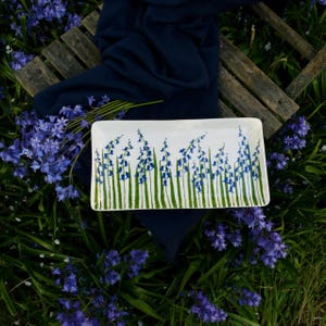 May include: A white rectangular ceramic serving dish with a blue and green floral design. The dish is decorated with a row of blue bellflowers with green stems and leaves.