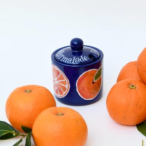 May include: A blue ceramic jar with a lid and a white inscription that reads "Marmalade". The jar is decorated with orange slices. There are several whole oranges in the background.