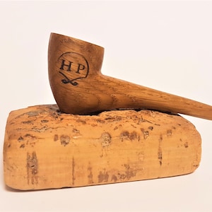 May include: A wooden pipe with a brown finish and the initials "HP" engraved on the bowl. The pipe is resting on a piece of cork.