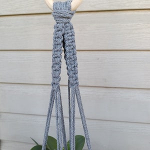 May include: Gray macrame plant hanger with a wooden ring at the top. The hanger is made of thick cotton cord and has a simple design.