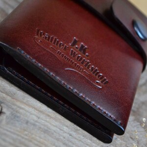 May include: A brown leather wallet with a black stitched edge and a flap closure. The wallet has the text "J.K. Leather Workshop - Handmade" embossed on the front.