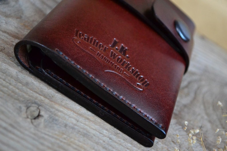 May include: A brown leather wallet with a black stitched edge and a flap closure. The wallet has the text "J.K. Leather Workshop - Handmade" embossed on the front.