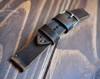 May include: A dark brown leather watch strap with stitching detail and a silver buckle. The strap is on a wooden surface.