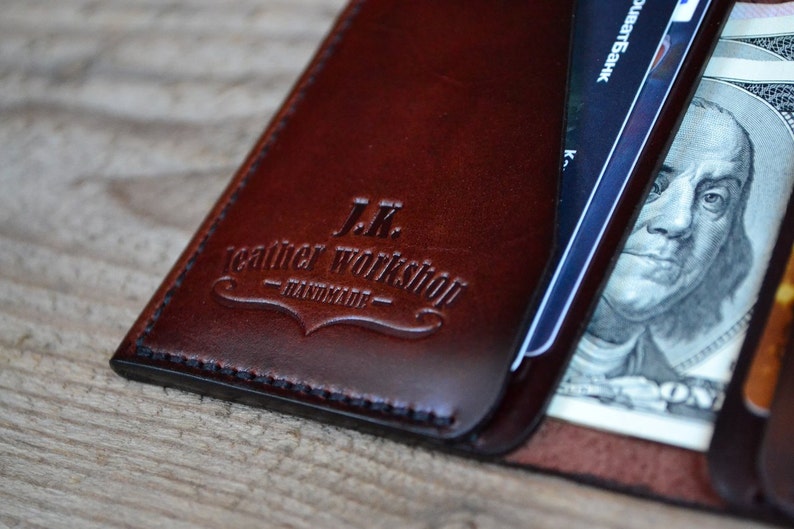 May include: A brown leather wallet with the text "J.K. Leather Workshop - Handmade" embossed on the front. The wallet is open and shows a credit card and a dollar bill.