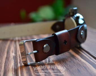 May include: A brown leather watch strap with a silver buckle and a metal button. The strap is handmade and has the text "J.K. leather workshop handmade" on it.