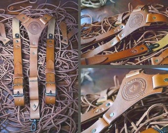 May include: Handmade leather suspenders in shades of tan and brown. The suspenders feature a Y-back design with adjustable straps, metal hardware, and a decorative emblem. The leather is embossed with a circular design and text.