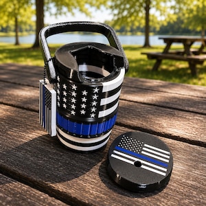 May include: A black, white, and blue beverage cooler with a handle, featuring a thin blue line flag design. A matching circular lid sits nearby, and a small rectangular tag with the same flag pattern hangs from the handle by a black chain.