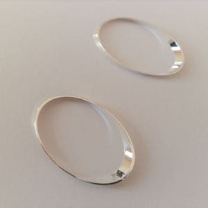 Puede incluir: Dos componentes de pendientes ovalados de plata pulida. Los pendientes tienen una superficie lisa y reflectante y están colocados sobre un fondo claro. El diseño es simple y elegante, adecuado para la fabricación de joyas.