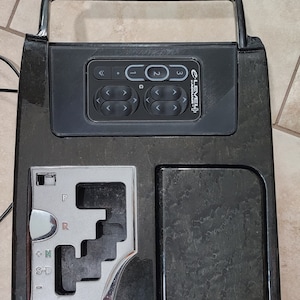 May include: Black car console with a silver gear shifter and a black control panel with buttons labeled "<" "." "1" "2" "3" and "E-LEVEL+  PRECISION".