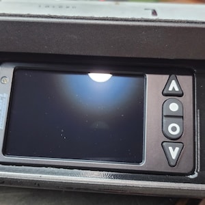 May include: A black dashboard component with a digital display screen and control buttons. The screen is dark, and the buttons are labeled with up, down, and center symbols. The component is set into a larger black housing.