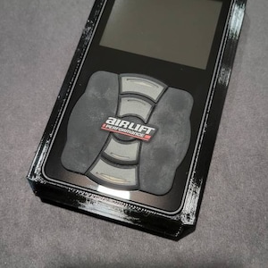 May include: A black electronic device with a screen and buttons, featuring the "AIRLIFT PERFORMANCE" logo. The device has a rectangular shape with a textured frame and a dark gray background.