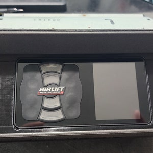 May include: A black air suspension control system with a digital display and a circular control panel. The control panel has a red "AirLift Performance" logo. The system is mounted in a black plastic housing.