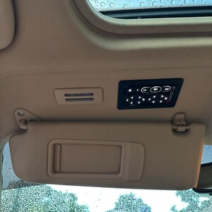 May include: A beige car interior with a sunroof, a sun visor, and a black control panel with buttons. The sunroof is covered in raindrops.