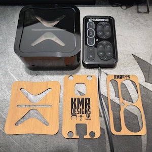 May include: A black control panel with a clear plastic cover and a black button panel labeled "E-Level" with buttons numbered 1, 2, and 3. The control panel is connected to a black cord. Three wooden cutouts with the text "KMR Designs" are shown. The cutouts are designed to hold the control panel and cord.