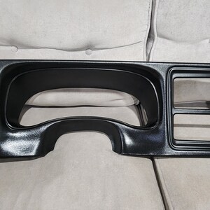 May include: Black plastic dashboard panel for a car. The panel has a large circular cutout for the speedometer and several smaller rectangular cutouts for other gauges.