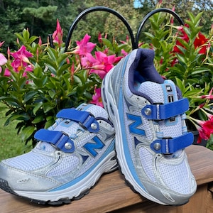 Puede incluir: Un par de zapatillas deportivas blancas con detalles en azul y plata. Las zapatillas tienen correas de velcro con detalles en azul y el logotipo de New Balance en el lateral.