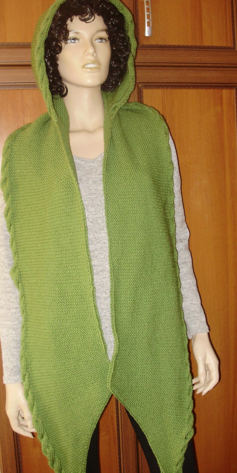 May include: A green knitted hooded scarf with a long, pointed design. The scarf is made of a soft, textured yarn and has a loose, flowing fit.