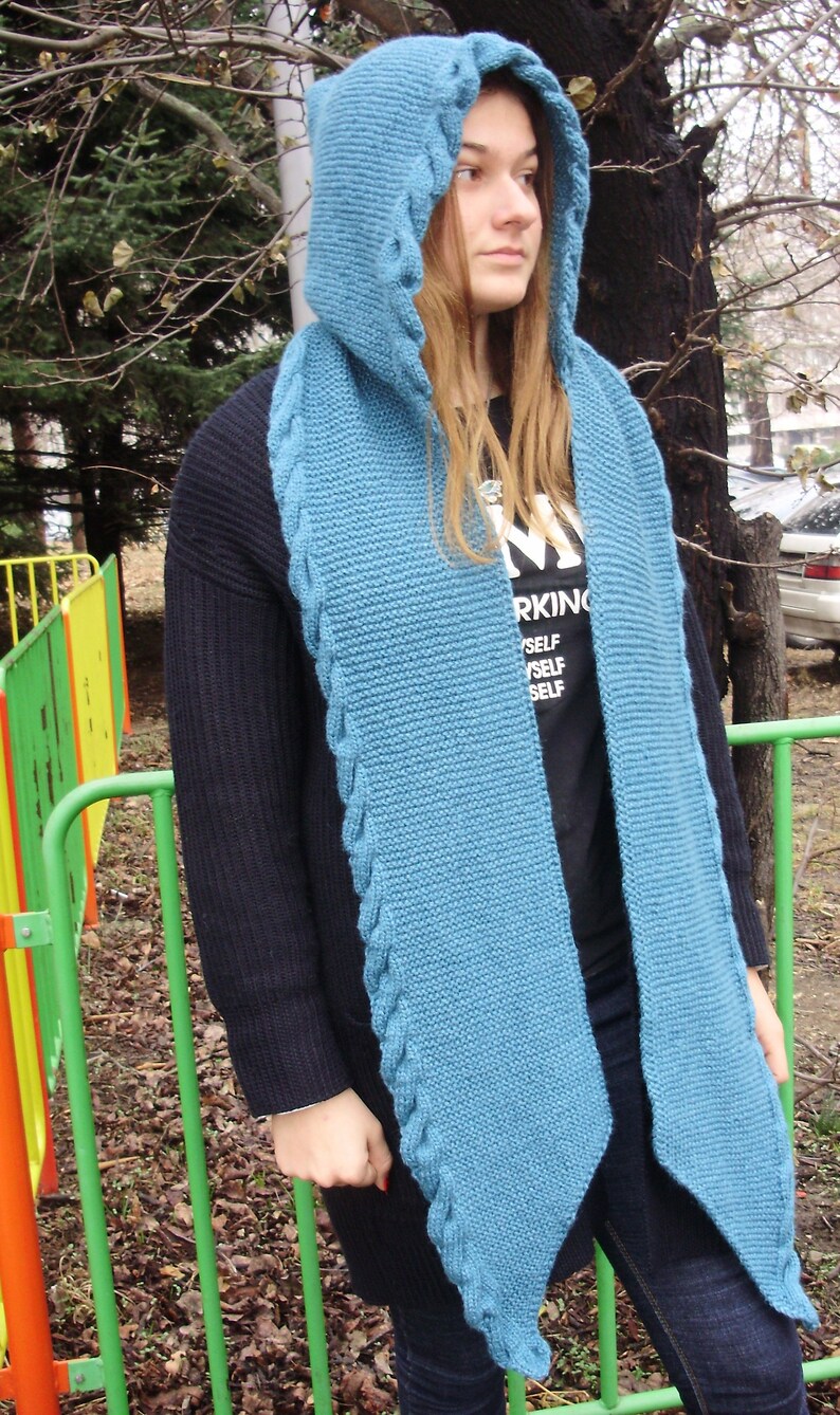 May include: A blue knitted hooded scarf with a cable knit pattern. The scarf is worn by a person with long brown hair and a black shirt that says "Working Myself Self".