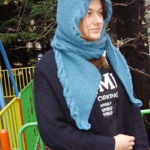 May include: A teal blue knitted hooded scarf with a cable knit pattern. The scarf is worn by a person wearing a black sweater and a black t-shirt with the text "Working Myself Myself Myself".