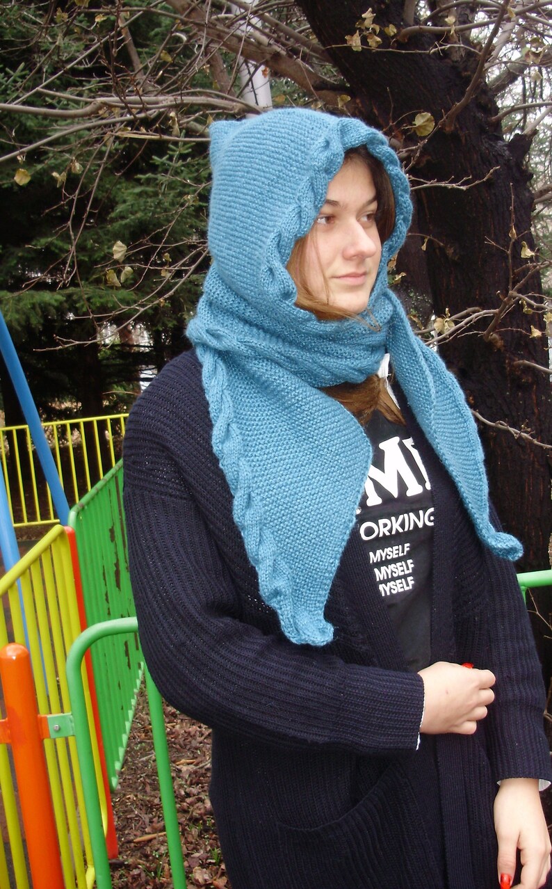 May include: A teal blue knitted hooded scarf with a cable knit pattern. The scarf is worn by a person wearing a black sweater and a black t-shirt with the text "Working Myself Myself Myself".
