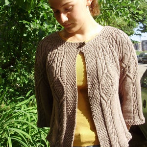 Peut inclure: Un cardigan en tricot marron avec un motif en câble et une fermeture à bouton unique. Le cardigan est porté sur une chemise jaune et un jean bleu.