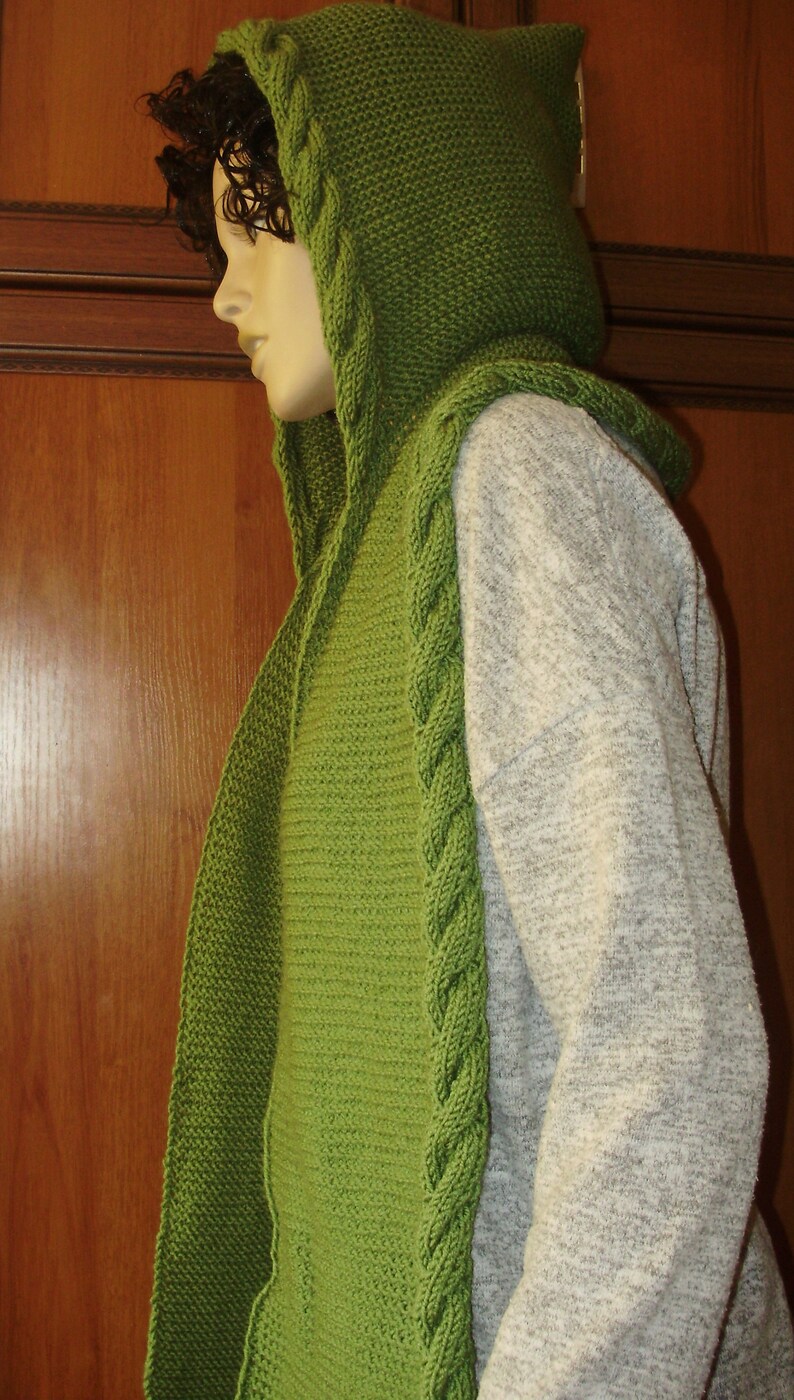 May include: A green knitted hooded scarf with a cable knit pattern. The scarf is worn by a person with dark hair.