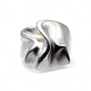 Könnte beinhalten: Ein silberner Ring mit einem einzigartigen, strukturierten Design. Der Ring hat eine fließende, organische Form mit einer leicht rauen Oberfläche. Das Metall scheint poliert zu sein und reflektiert das Licht. Der Ring ist vor einem schlichten weißen Hintergrund platziert.