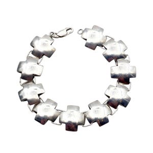 Könnte beinhalten: Ein silberfarbenes Armband mit einer Reihe von kreuzförmigen Gliedern. Das Armband hat einen Karabinerverschluss. Die Kreuze sind flach und haben eine leicht strukturierte Oberfläche. Der Schmuck wird vor weißem Hintergrund präsentiert.