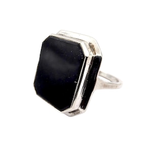 Könnte beinhalten: Ein silberner Ring mit einem großen, rechteckigen schwarzen Stein. Der Stein ist in einer silbernen Lünette mit geometrischem Design gefasst. Der Ring hat ein schlichtes Band.