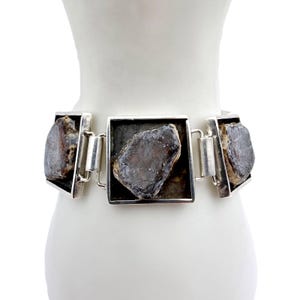 Könnte beinhalten: Ein silbernes Armband mit drei quadratischen Gliedern, die jeweils einen dunklen, unregelmäßig geformten Stein enthalten. Die Steine weisen ein gesprenkeltes Muster in Braun- und Grautönen auf. Das Armband wird auf einer weißen Schaufensterpuppe präsentiert.