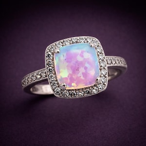 Könnte beinhalten: Silberner Ring mit einem quadratischen, schillernden Opal-Mittelstein in Rosa, Blau und Gelb. Der Opal ist von einem Kranz aus kleinen, klaren Edelsteinen umgeben, mit weiteren Steinen am Ringband. Ein schönes Schmuckstück.
