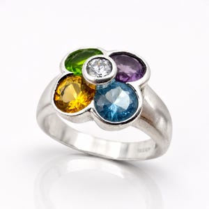 Könnte beinhalten: Silberring mit einem Blumenmuster, besetzt mit sechs farbigen Edelsteinen. Die Edelsteine umfassen gelbe, grüne, violette und blaue Steine, mit einem klaren Stein in der Mitte. Auf weißem Hintergrund.