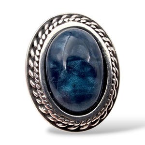 Könnte beinhalten: Ovaler Ring mit einem dunkelblauen, schimmernden Stein. Der Stein ist in einen silberfarbenen Metallrahmen mit einem geflochtenen Seildesign am Rand gefasst. Auf weißem Hintergrund.