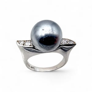 Può includere: Un anello d'argento con una grande perla grigia come elemento centrale. La fascia dell'anello è adornata da piccole pietre preziose trasparenti su entrambi i lati della perla, aggiungendo brillantezza. L'anello ha un design moderno ed elegante.