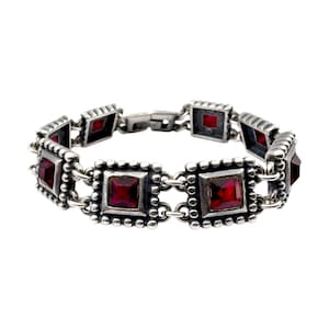 Könnte beinhalten: Silberarmband mit quadratischen, tiefroten Edelsteinen, die in verzierten, mit Perlen besetzten Rahmen gefasst sind. Das Armband hat ein klassisches Design mit einem sicheren Verschluss. Die Edelsteine haben eine lebendige Farbe.