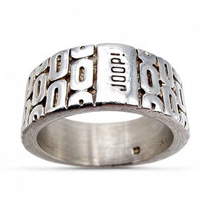 Könnte beinhalten: Ein silberfarbener Ring mit einem sich wiederholenden Muster aus rechteckigen Aussparungen. Das Wort "JOOP!" ist vertikal auf einem erhabenen rechteckigen Abschnitt dargestellt. Der Ring hat ein gebürstetes Metall-Finish und ein breites Band.