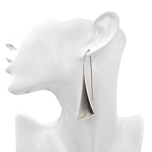 May include: A pair of silver drop earrings. Each earring has a long, thin hook that leads to a triangular, flared pendant. The earrings are shown on a white mannequin head against a white backdrop.