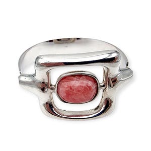 Könnte beinhalten: Ein silberfarbenes Armband mit rechteckigem Design und einem ovalen, rosafarbenen Edelstein in der Mitte. Das Armband hat einen modernen, geometrischen Stil mit poliertem Finish. Der Edelstein hat ein gesprenkeltes Aussehen.