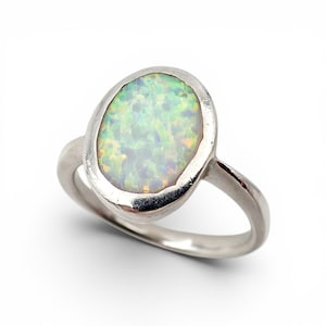 Könnte beinhalten: Ein silberfarbener Ring mit einem großen, ovalen Cabochon-Stein, der eine milchig-weiße Basis mit irisierenden grünen, blauen und gelben Lichtreflexen aufweist. Die Metallfassung hat eine glatte, polierte Oberfläche und eine schlichte Ringschiene.