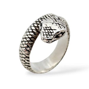 Könnte beinhalten: Ein silberfarbener Ring in Form einer Schlange. Der Schlangenkopf ist mit strukturierten Schuppen und kleinen Augen versehen. Der Ring umschlingt sich, wobei der Schlangenkopf nach außen zeigt. Der Ring besteht aus glänzendem Metall.