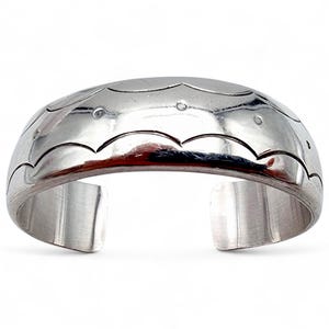 Könnte beinhalten: Ein silbernes Armband mit einem wellenförmigen Design und kleinen Punktakzenten. Das offene Manschettendesign ermöglicht ein einfaches Tragen. Das Armband besteht aus glänzendem, reflektierendem Metall.