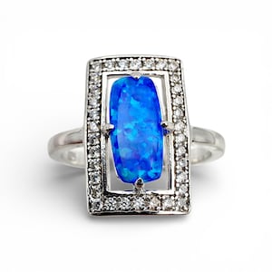 Könnte beinhalten: Silberner Ring mit einem rechteckigen, leuchtend blauen Opal. Der Opal ist von einer Fassung aus kleinen, klaren Edelsteinen umgeben. Der Ring hat ein schlichtes, poliertes Silberband.