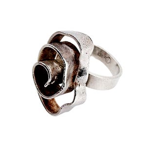 May include: A silver ring with an unusual, abstract design. The ring has a swirling, textured centrepiece, like a stylised flower or spiral. The band is smooth, with a hallmark visible. The ring is set against a white background. The ring is a statement piece.