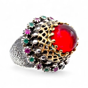 Könnte beinhalten: Ein auffälliger Ring mit einem großen, leuchtend roten Edelstein im Mittelpunkt. Der Edelstein ist in eine goldene Filigranfassung gesetzt und mit kleineren grünen und rosa Steinen verziert. Der Ring hat ein strukturiertes Silberband.