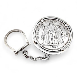 Könnte beinhalten: Silberner Schlüsselanhänger mit einer Münze, auf der die Worte "LIBERTE EGALITE FRATERNITE" und eine Darstellung von drei Figuren zu sehen sind. Der Schlüsselanhänger hat einen Ring und eine Kette.
