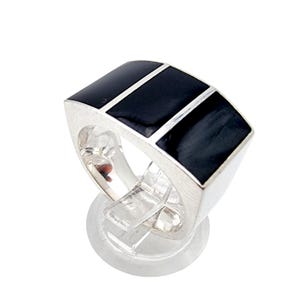 May include: A silver ring with a rectangular face featuring three black enamel insets separated by thin silver lines. The ring is displayed on a clear, round stand, showcasing its geometric design and polished finish. The ring's design is modern and bold.