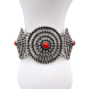 Könnte beinhalten: Silberfarbenes Armband mit einem zentralen kreisförmigen Design und roten Steinakzenten. Das Armband hat aufwendige Details mit kleinen, runden Verzierungen. Es wird auf einer weißen Oberfläche präsentiert. Ein Schmuckstück.